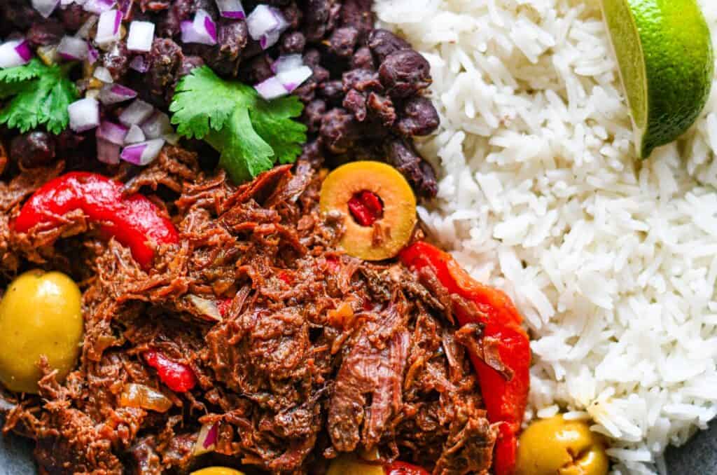 Cuban Ropa Vieja con Venado with black beans and rice and garnished with red onion and cilantro.