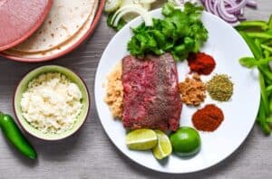 Deer Steak Fajita ingredients including yellow onions, red onions, bell peppers, minced garlic, cilantro, cumin, smoked paprika, chili powder, oregano, queso fresco, limes, and tortillas.