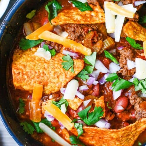 Deer Steak Chili with beef broth, kidney beans, garlic, celery, and honey and garnished with cilantro, onions, cheese, and Doritos.