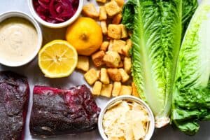 Venison Steak Caesar Salad ingredients including romaine lettuce, shaved Parmesan, Caesar dressing, croutons, lemons, pickled red onion, and venison steak